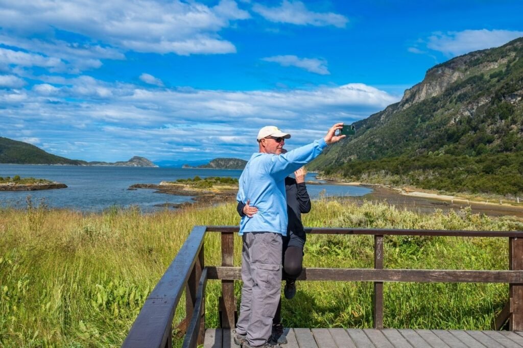 Turista sacándose una selfie en un mirador de madera frente a Bahía Lapataia, con islotes, pastizales y montañas al fondo en el Parque Nacional Tierra del Fuego