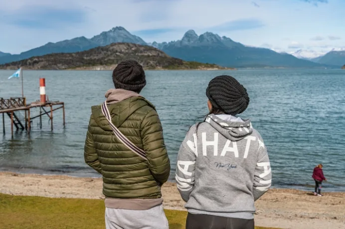 Dos turistas de espaldas contemplan las montañas y el agua cristalina de la Bahía Ensenada Zaratiegui, Ushuaia.