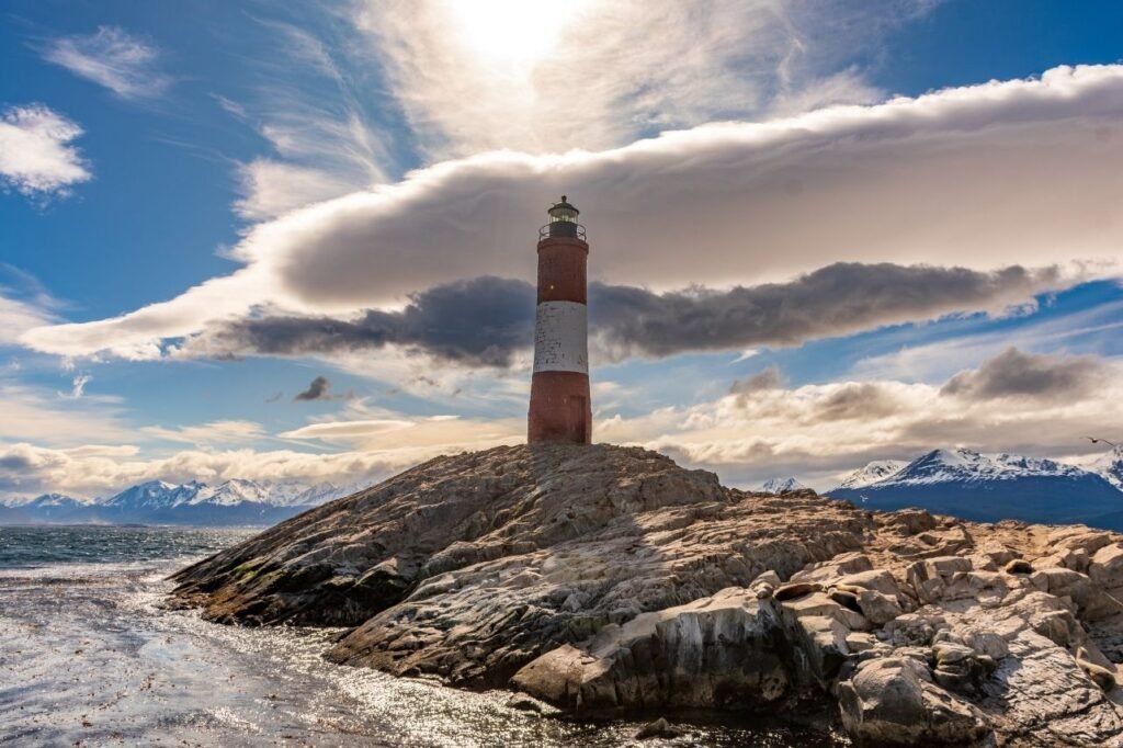 Faro Les Eclaireurs a contraluz sobre rocas con lobos marinos, con nubes dramáticas y montañas nevadas en el Canal Beagle, Tierra del Fuego