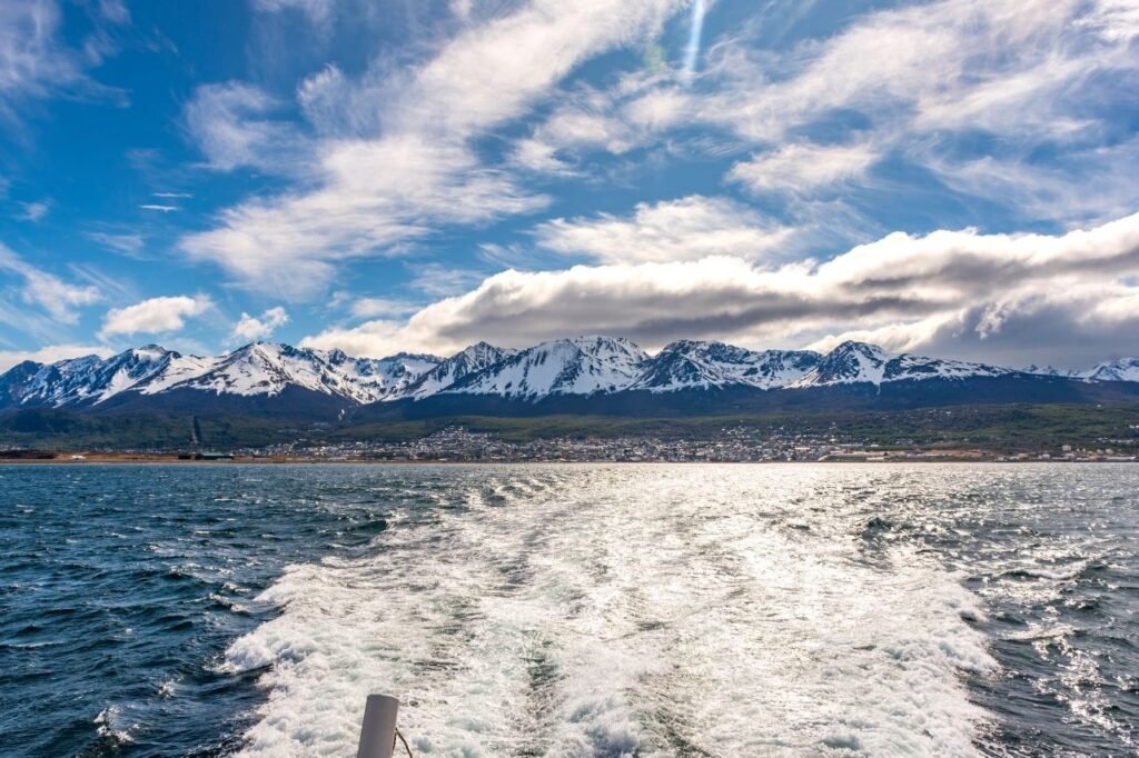 Vista de la ciudad de Ushuaia al pie de montañas nevadas desde el catamarán, con la estela de espuma sobre el Canal Beagle