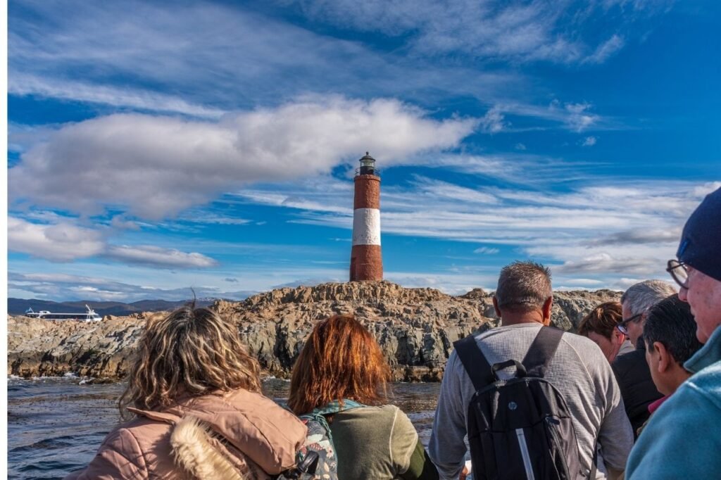 Turistas de espaldas observando el Faro Les Eclaireurs de franjas rojas y blancas sobre un islote rocoso en el Canal Beagle, Ushuaia