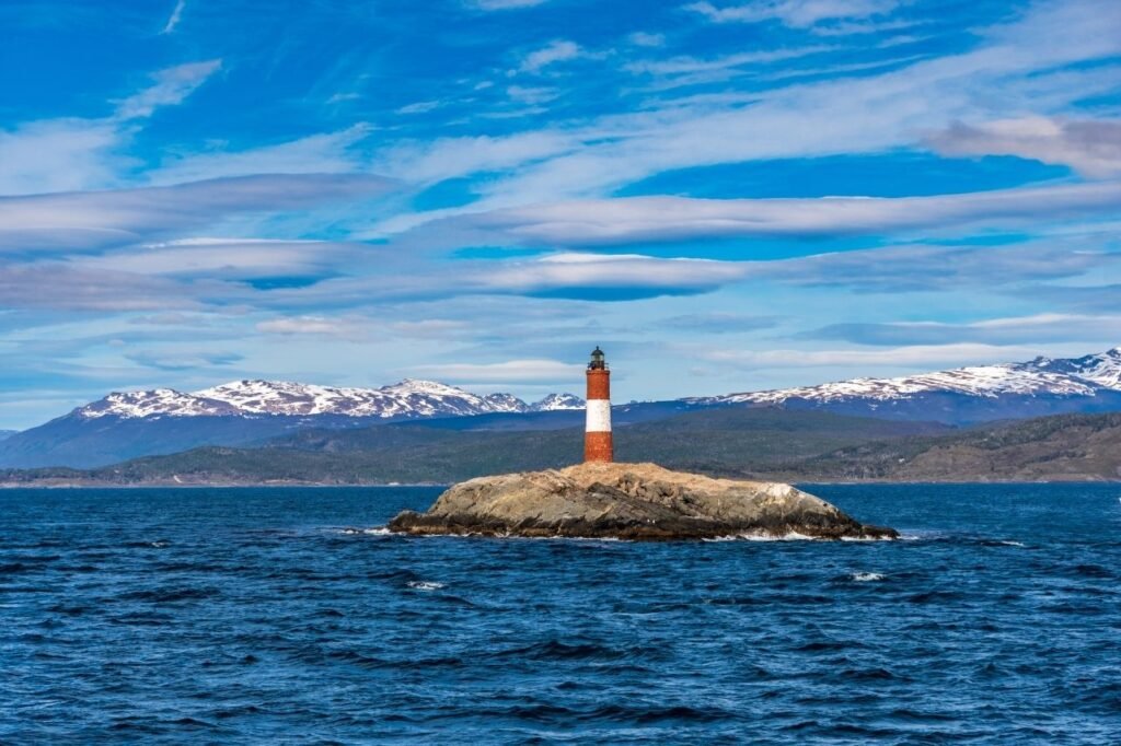 Faro Les Eclaireurs de franjas rojas y blancas sobre un islote rocoso en aguas azules del Canal Beagle, con montañas nevadas al fondo en Ushuaia