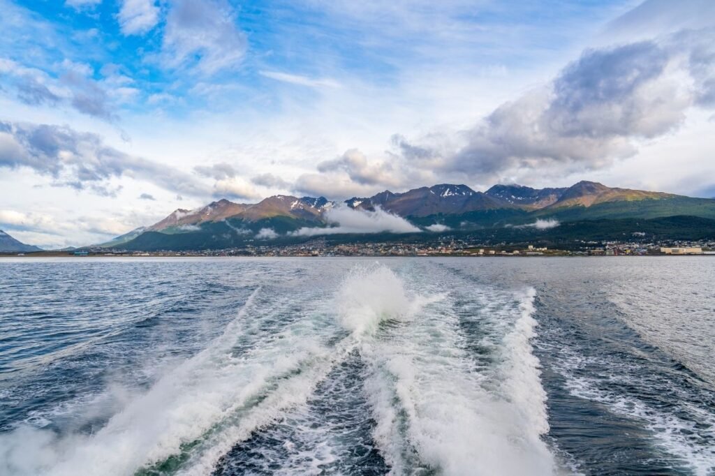 Vista de la ciudad de Ushuaia al pie de las montañas, con nubes bajas y la estela de espuma del catamarán sobre el Canal Beagle