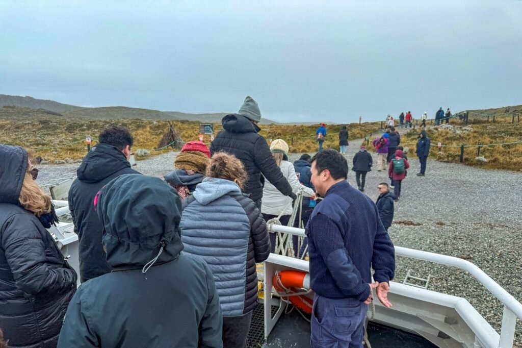 Pasajeros bajando del catamarán a una isla del Canal Beagle, con un tripulante ayudando y un sendero entre pastizales en Tierra del Fuego