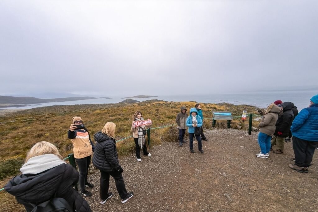 Turistas sacándose fotos en un mirador de una isla del Canal Beagle, con una mujer sosteniendo un cartel con el texto #Ushuaia y el canal con bruma al fondo