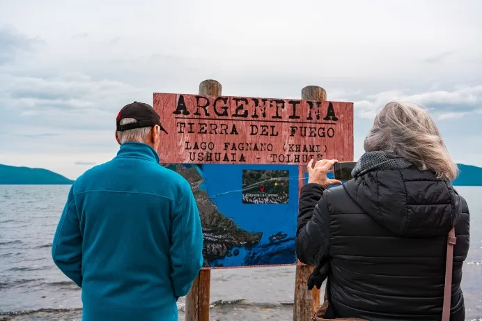 Una pareja de turistas de Aswalek Expediciones se toma una foto y lee un cartel informativo en la orilla de piedras del lago Fagnano en Tierra del Fuego.