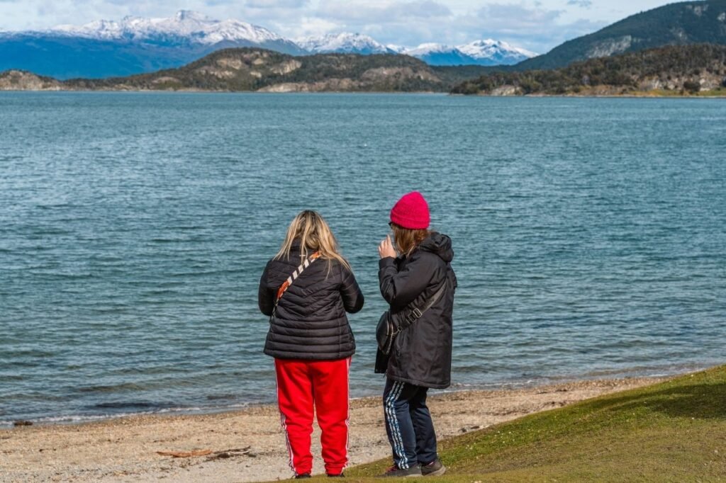 Dos visitantes de espaldas frente a las aguas azules de la bahía Ensenada, con montañas nevadas al fondo en el Parque Nacional Tierra del Fuego