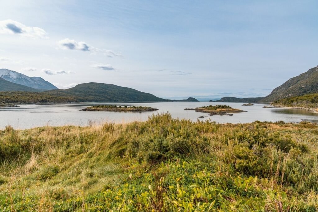 Bahía Lapataia, de aguas calmas con pequeños islotes, pastizales en primer plano y montañas con nieve al fondo en el Parque Nacional Tierra del Fuego
