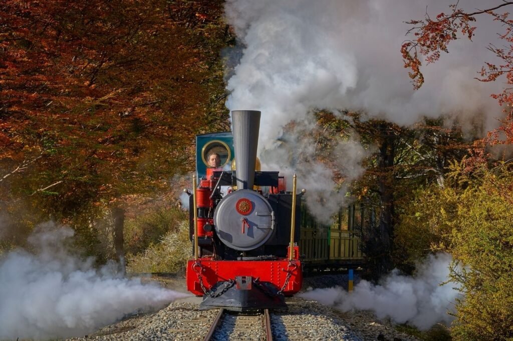 Locomotora a vapor roja del Tren del Fin del Mundo de frente, rodeada de vapor y árboles otoñales en Ushuaia