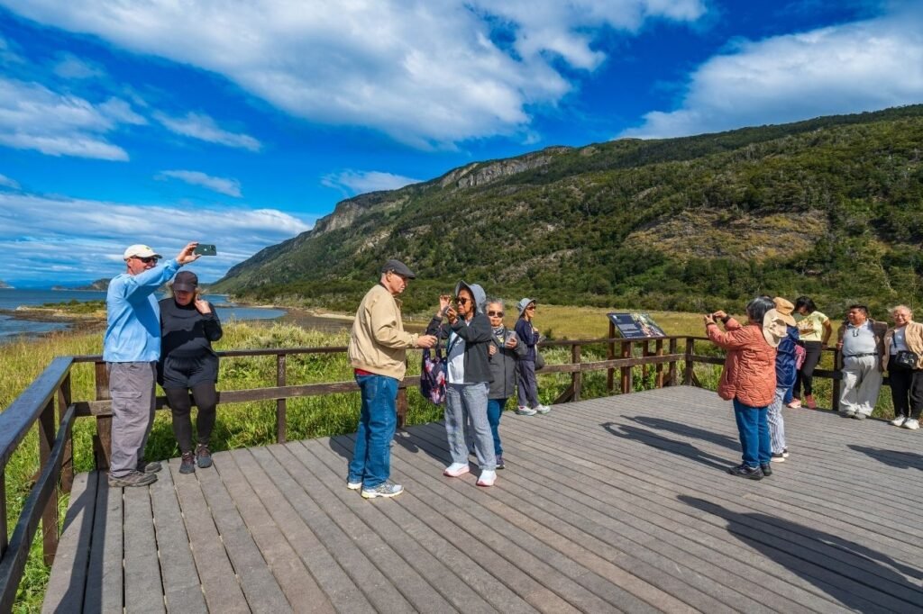 Turistas sacando fotos en una plataforma de madera con baranda y cartel informativo, con vista a Bahía Lapataia y montañas boscosas en el Parque Nacional Tierra del Fuego
