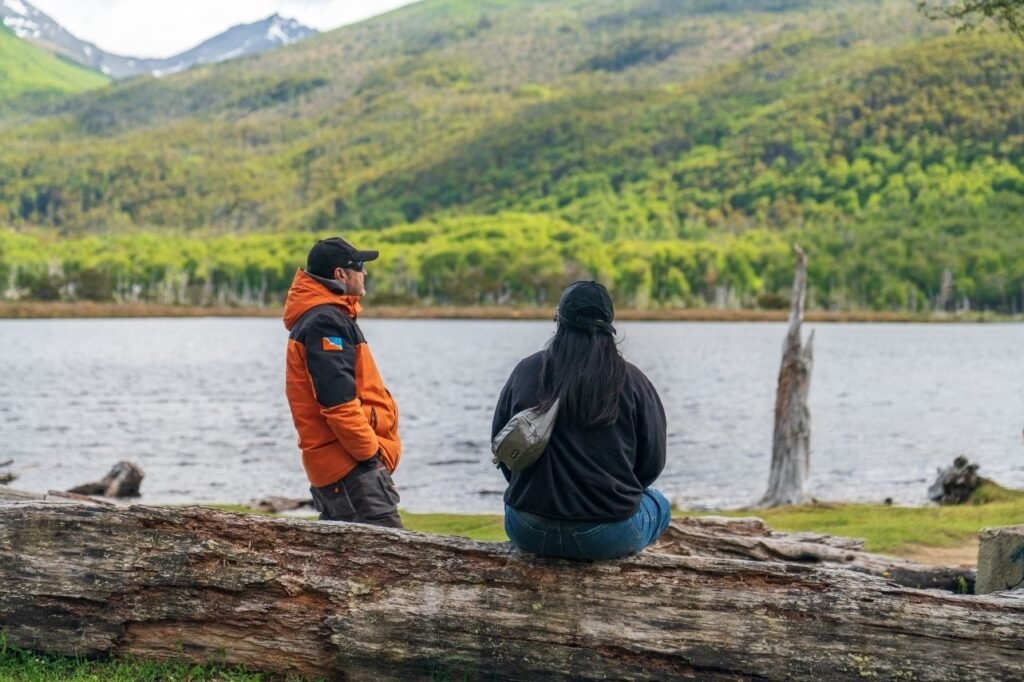 Guía y turista conversando sentados sobre un tronco frente a Laguna Victoria y el bosque, Tierra del Fuego