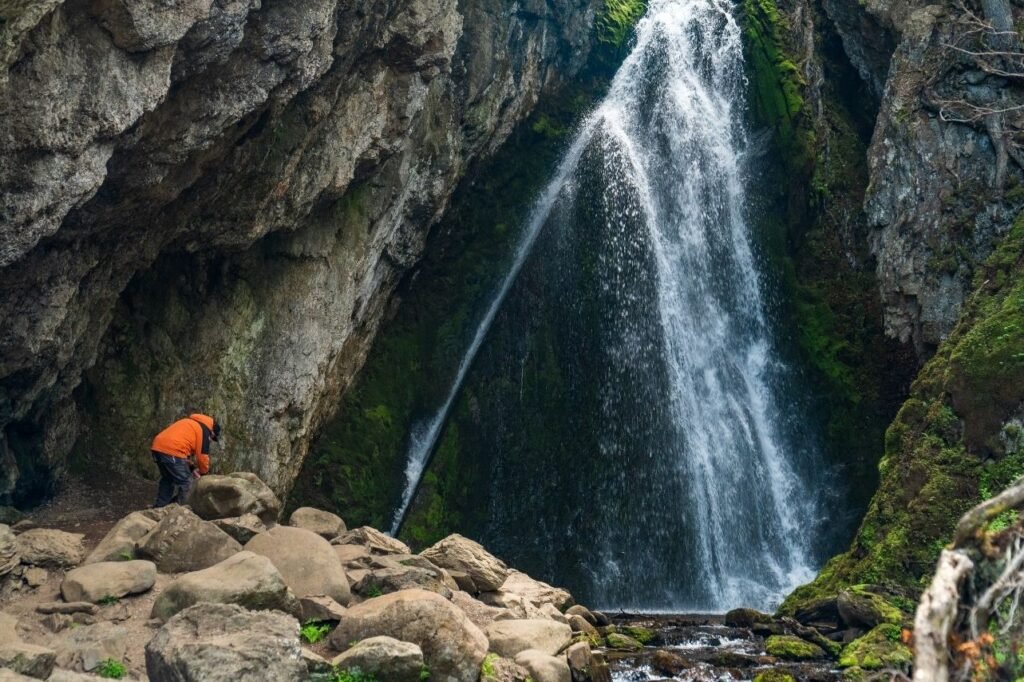 Excursionista con campera naranja frente a la Cascada del Duende, entre paredes de roca cubiertas de musgo