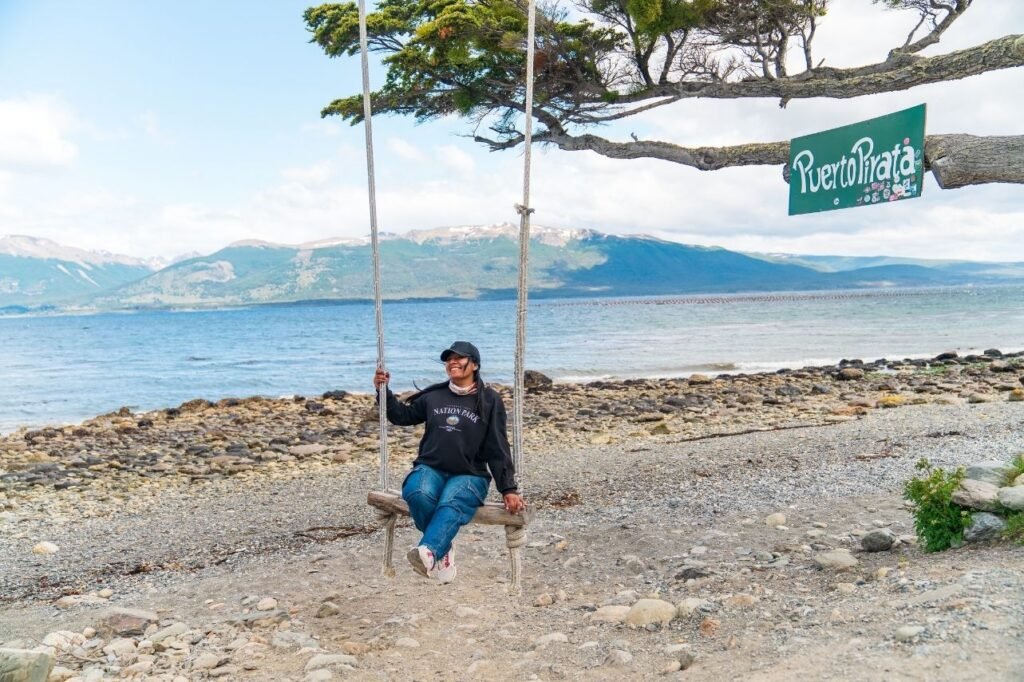 Turista sentada en el columpio junto al cartel de Puerto Pirata, con el Canal Beagle y montañas de fondo