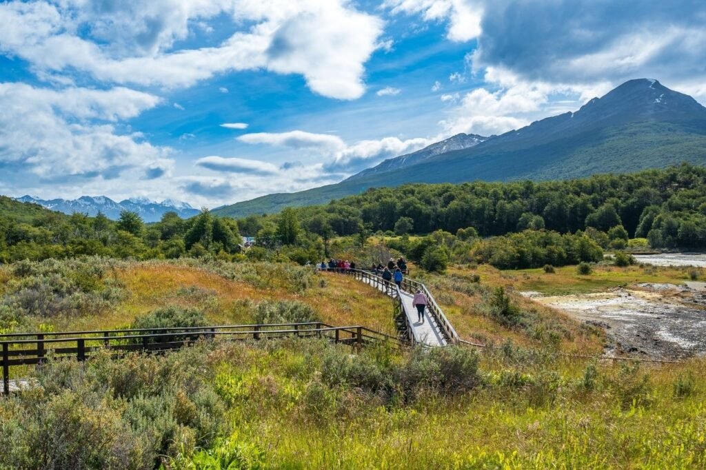 Grupo de visitantes recorriendo una pasarela de madera entre pastizales y bosque, con montañas y cielo con nubes en Bahía Lapataia, Parque Nacional Tierra del Fuego