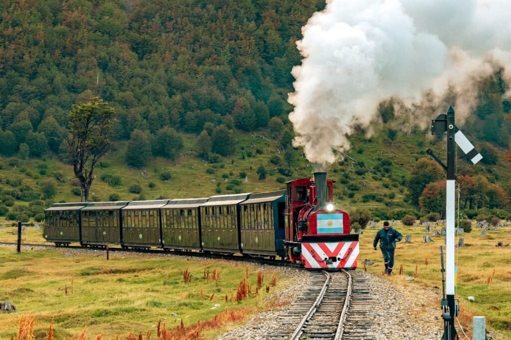 Locomotora a vapor roja con bandera argentina y vagones verdes avanzando por la vía entre bosque otoñal y pastizales en Tierra del Fuego