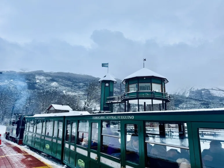 Primer plano de los vagones verdes del Tren del Fin del Mundo y la torre de control de madera bajo la nieve en Ushuaia.