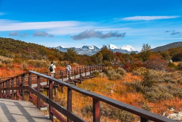 Dos excursionistas caminando por una pasarela de madera en el Parque Nacional Tierra del Fuego, rodeados de colores otoñales y montañas nevadas.