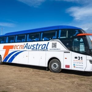 Autobús de larga distancia de la empresa TecniAustral, modelo Mercedes-Benz, estacionado en Ushuaia. Vista lateral derecha mostrando la marca y colores blanco, naranja y azul.