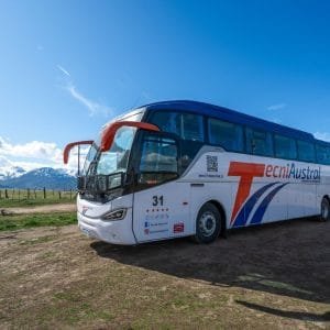 Frente diagonal del autobús TecniAustral en Ushuaia, destacando los espejos naranja, el logo Mercedes-Benz y la cordillera nevada de fondo.