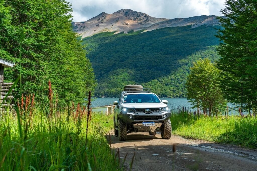 Camioneta 4x4 blanca con neumático de auxilio en el techo avanzando por un camino de tierra entre bosque, con el lago Escondido y una montaña al fondo en Tierra del Fuego.