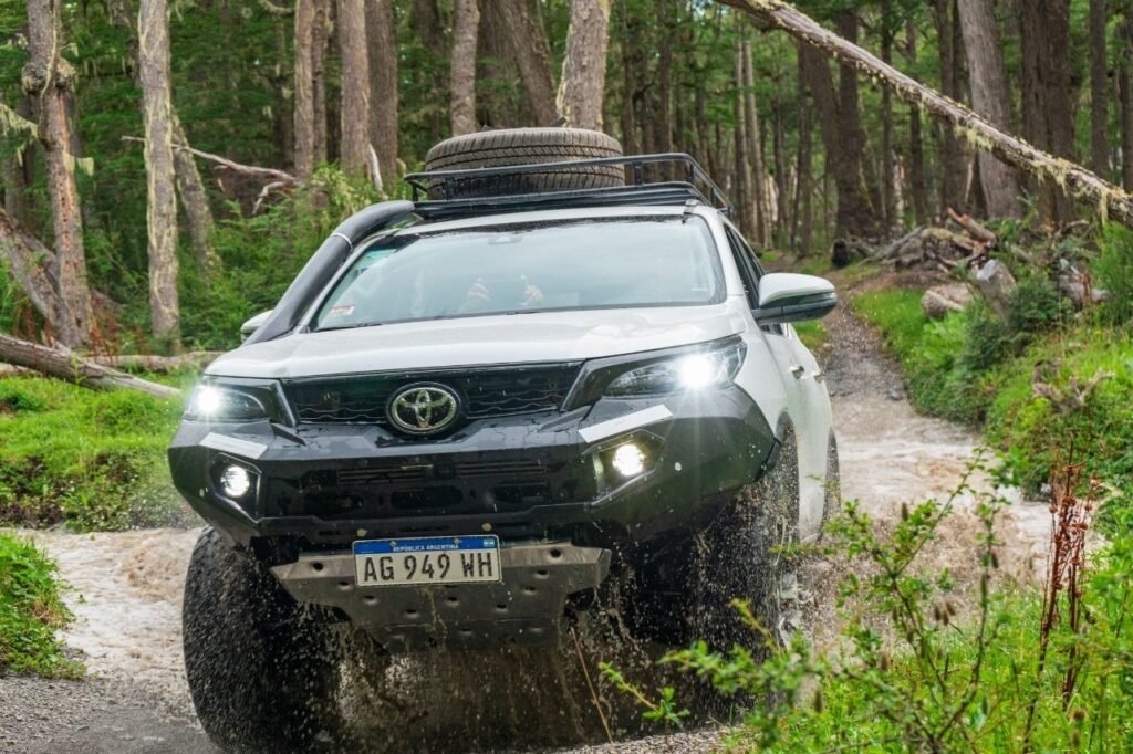 Camioneta 4x4 Toyota con snorkel y neumático de auxilio en el techo cruzando un arroyo con barro y agua salpicando dentro del bosque de Tierra del Fuego.