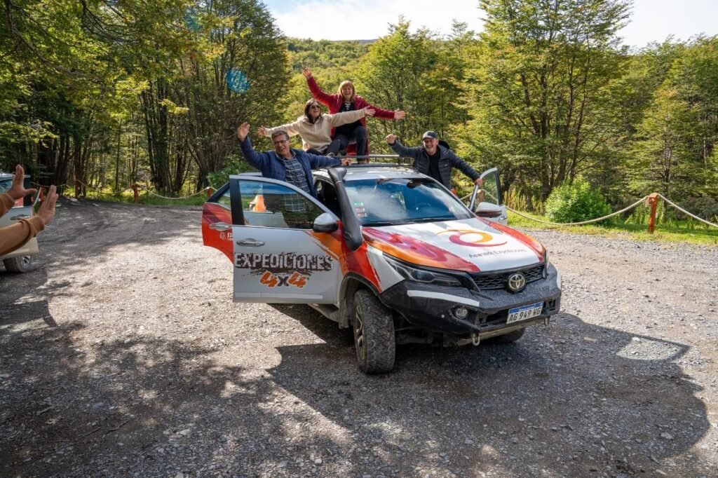 Cuatro turistas con los brazos abiertos asomados sobre una camioneta 4x4 de Expediciones 4x4 en un camino de ripio rodeado de bosque en Tierra del Fuego.