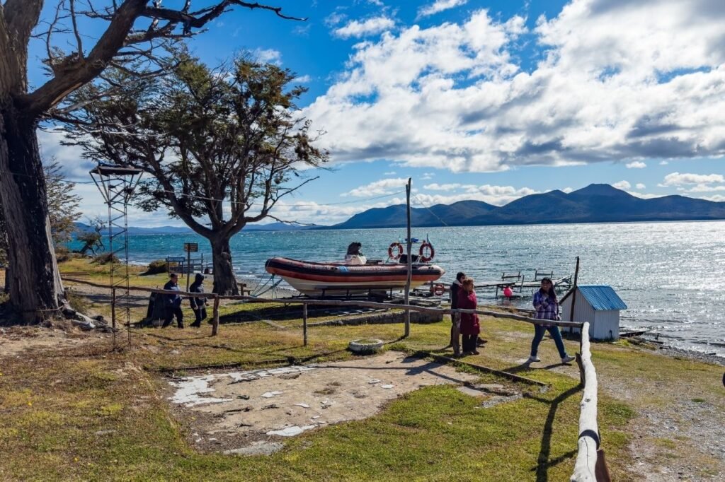 Orilla del Lago Fagnano con una lancha inflable sobre la costa, un muelle de madera, una casilla azul y visitantes caminando en Tierra del Fuego