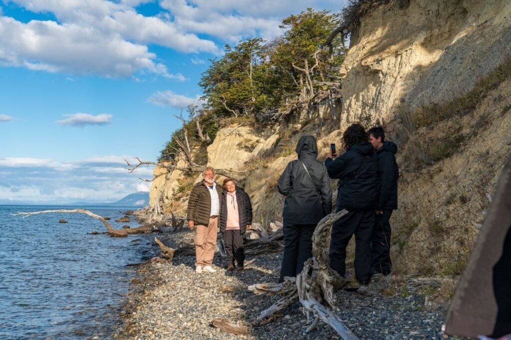 Grupo de turistas sacando fotos en la costa de piedras del Lago Fagnano, junto a troncos secos y una barranca con bosque en Tierra del Fuego.