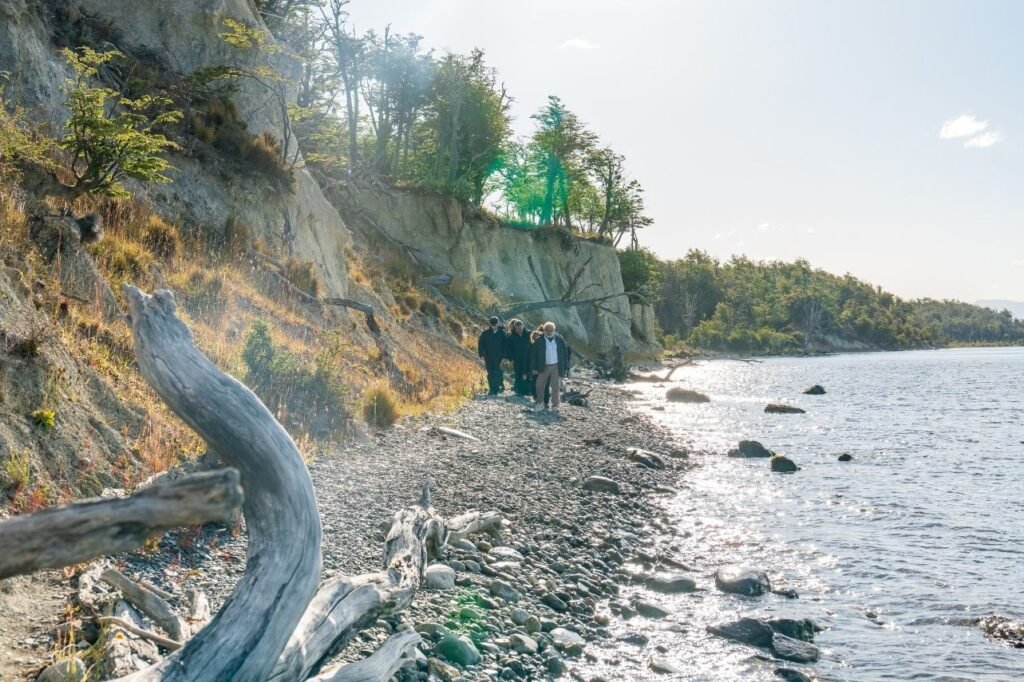 Personas caminando por la orilla de piedras del Lago Fagnano, al pie de una barranca con bosque y con troncos secos en primer plano en Tierra del Fuego.
