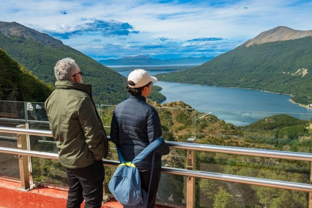 Dos turistas de espaldas observando el Lago Escondido y las montañas boscosas desde el Mirador Paso Garibaldi con baranda de vidrio en Tierra del Fuego