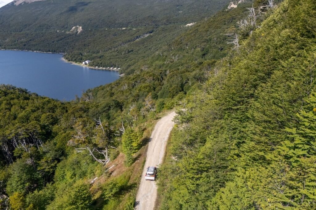 Vista aérea de una camioneta 4x4 por un camino de ripio entre bosque de lengas, con el Lago Escondido y unas cabañas en la costa al fondo en Tierra del Fuego.