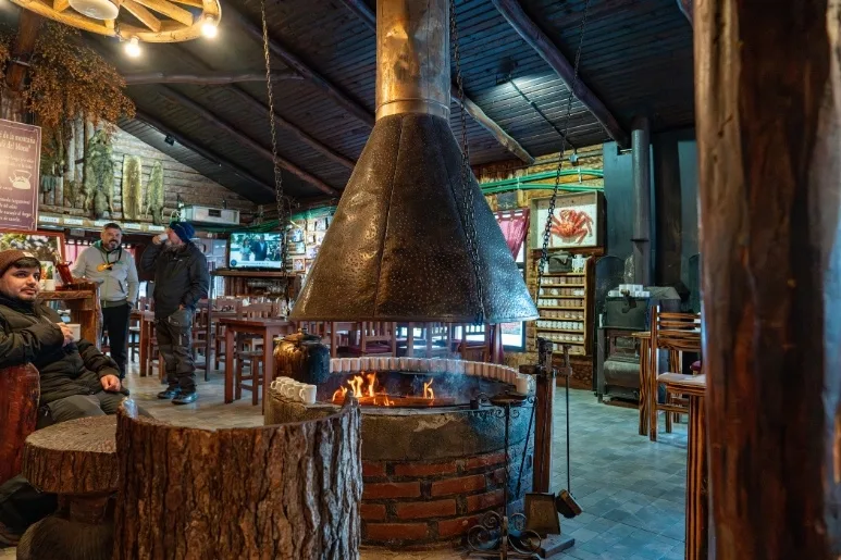 Interior rústico de refugio de montaña en Ushuaia con gran chimenea central de hierro y gente tomando calor. Ideal para excursiones de invierno.