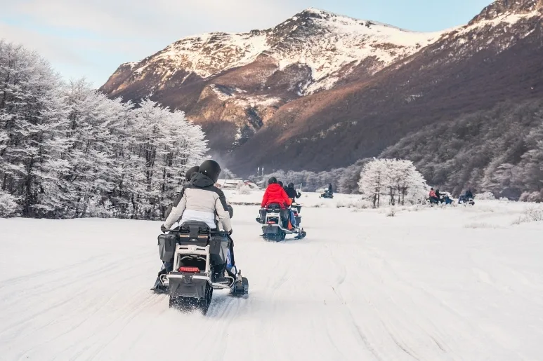 Pareja en moto de nieve siguiendo a guía en paisaje boscoso de Ushuaia.
