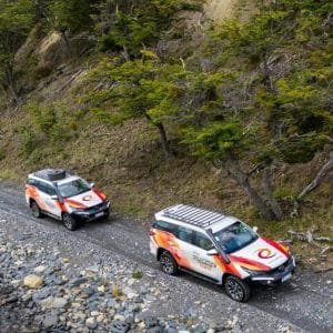 Dos camionetas 4x4 de Aswalek Expediciones circulando por la costa de piedras del Lago Fagnano, con el agua cristalina y el bosque de lengas bajo las montañas de Ushuaia al fondo.