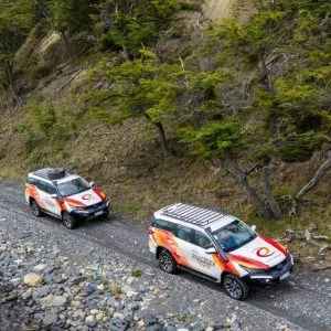 Dos camionetas 4x4 de Aswalek Expediciones circulando por la costa de piedras del Lago Fagnano, con el agua cristalina y el bosque de lengas bajo las montañas de Ushuaia al fondo.