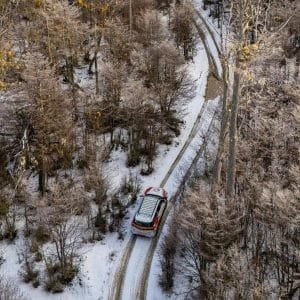 Una toma aérea cenital captura una camioneta blanca 4x4 conduciendo por un camino de tierra cubierto de nieve profunda, serpenteando a través de un denso bosque de lengas helado en un valle andino cerca de Ushuaia, bajo un cielo cubierto.