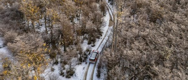 Una toma aérea cenital captura una camioneta blanca 4x4 conduciendo por un camino de tierra cubierto de nieve profunda, serpenteando a través de un denso bosque de lengas helado en un valle andino cerca de Ushuaia, bajo un cielo cubierto.