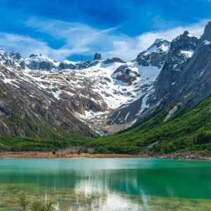 Paisaje panorámico de la Laguna Esmeralda en Ushuaia, mostrando sus aguas de color verde turquesa rodeadas de un bosque de lengas y las imponentes montañas nevadas de la Cordillera de los Andes bajo un cielo azul despejado.