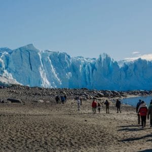 Un grupo de turistas camina por una playa de arena oscura hacia un enorme frente de glaciar azul bajo un cielo despejado.