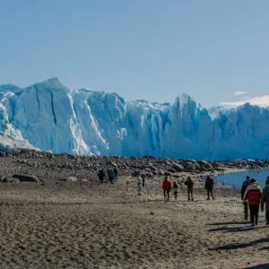Un grupo de turistas camina por una playa de arena oscura hacia un enorme frente de glaciar azul bajo un cielo despejado.