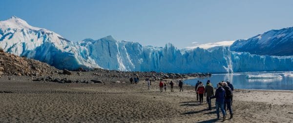 Un grupo de turistas camina por una playa de arena oscura hacia un enorme frente de glaciar azul bajo un cielo despejado.