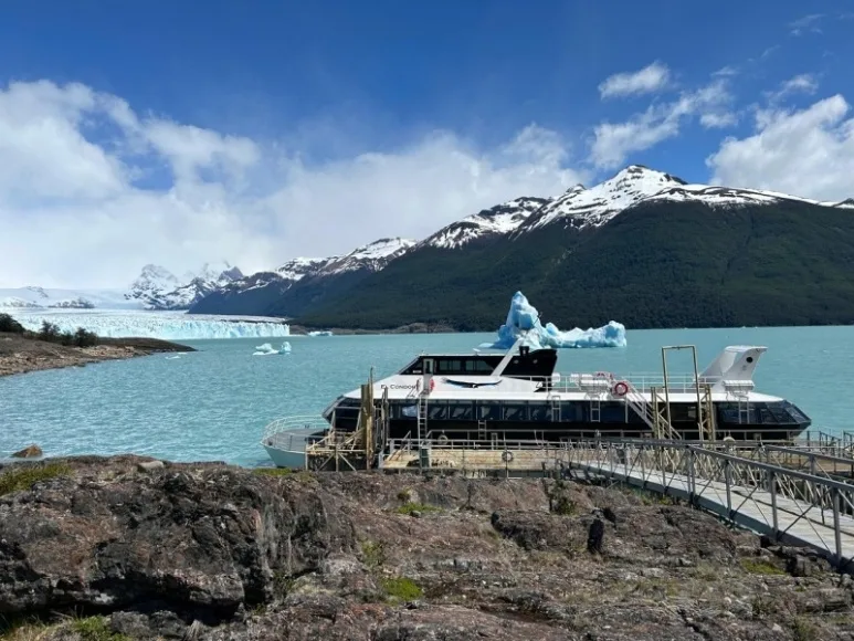 Un catamarán turístico atracado en un muelle frente al Lago Argentino con montañas nevadas y el glaciar Perito Moreno de fondo en la Patagonia.