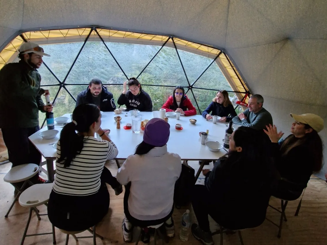 Grupo almorzando un guiso caliente en el domo geodésico con vista panorámica a la montaña nevada durante el Trekking Laguna Esmeralda.