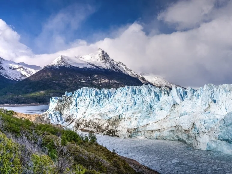 Una toma horizontal de la colosal pared de hielo azul agrietado del Glaciar Perito Moreno con montañas nevadas y dramáticas nubes al fondo.