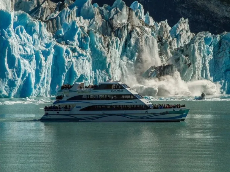 Un catamarán turístico lleno de gente en la cubierta navega cerca de la pared del Glaciar Perito Moreno mientras ocurre un desprendimiento de hielo con una nube de espuma.