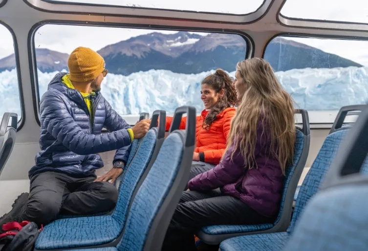 Grupo de turistas charlando y sonriendo dentro de un catamarán con vistas al glaciar.