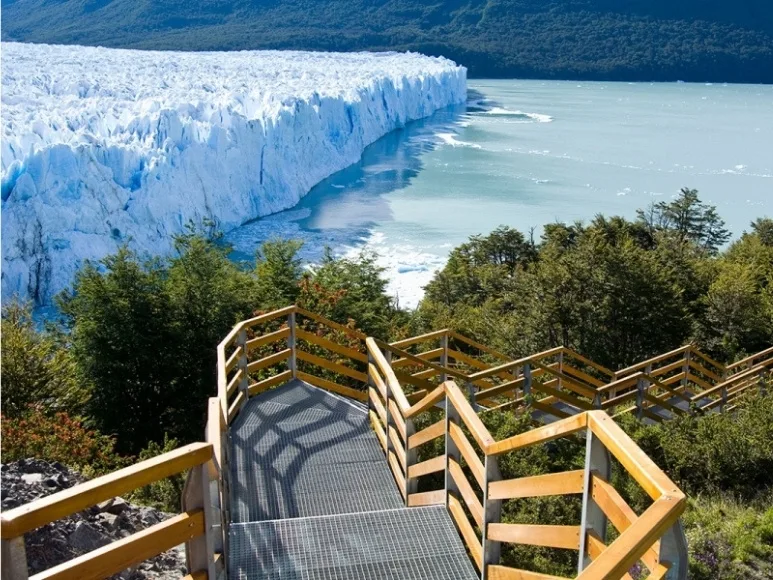 Una vista en ángulo alto desde unas pasarelas de madera y metal serpenteantes que ofrecen una panorámica del Glaciar Perito Moreno y el Lago Argentino.