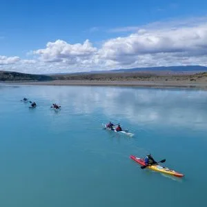Grupo de turistas en kayaks dobles y simples navegando en fila por las aguas tranquilas y turquesas del Lago Argentino bajo un cielo parcialmente nublado.