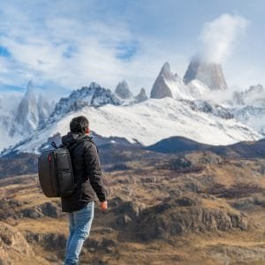 Un turista de espalda con mochila negra contemplando el imponente Cerro Fitz Roy nevado desde una cumbre rocosa sobre el valle.