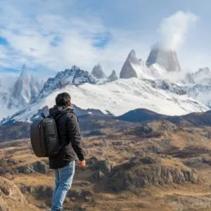 Un turista de espalda con mochila negra contemplando el imponente Cerro Fitz Roy nevado desde una cumbre rocosa sobre el valle.